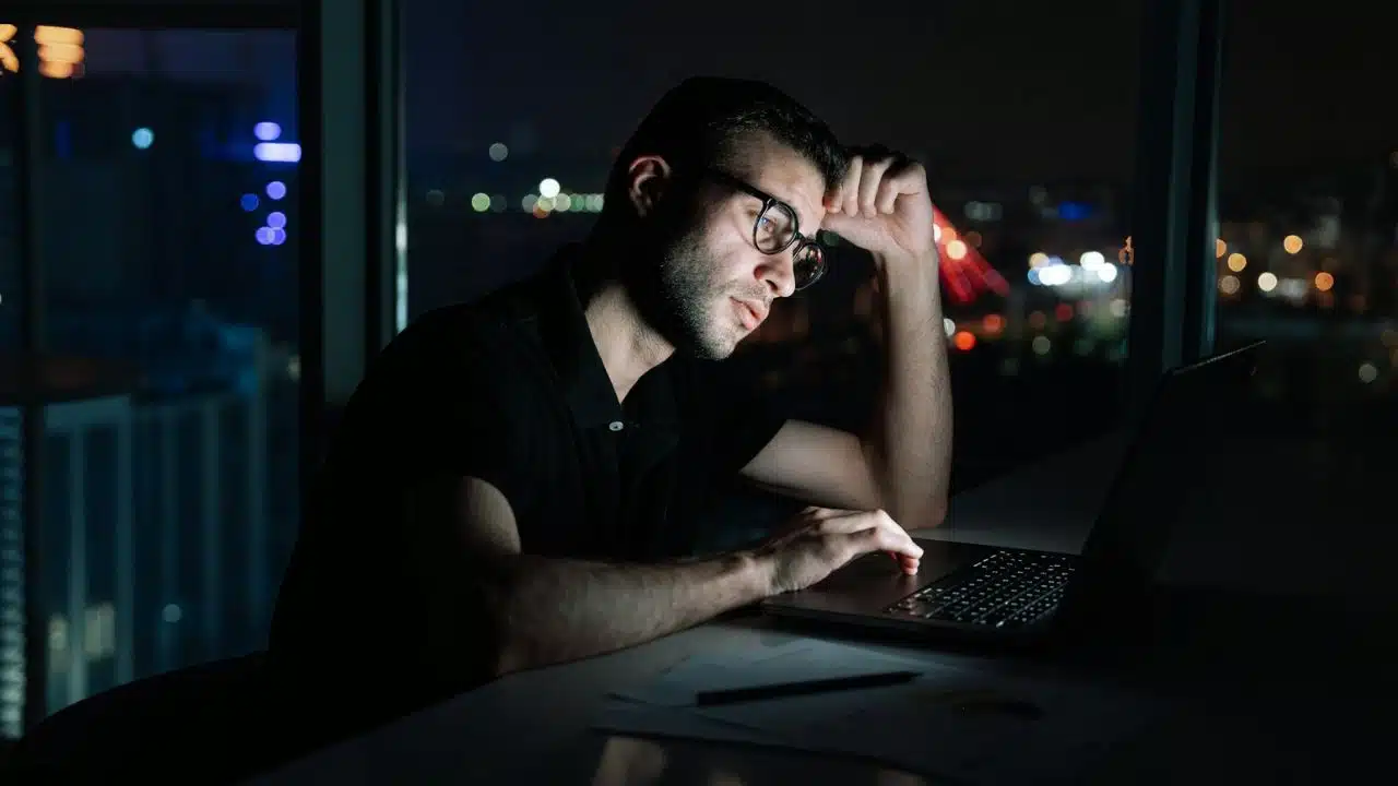 A man with glasses working on a laptop late at night in a dark room.