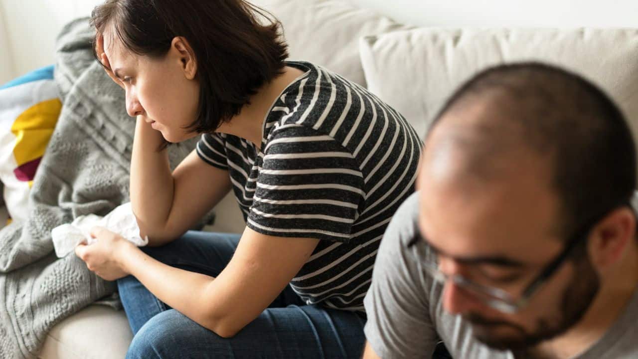 A woman looks upset on a couch, holding her head, with a man out of focus nearby.