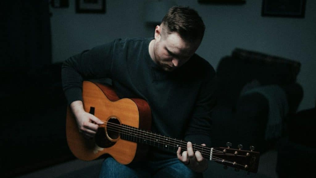 A man in a dark shirt sits in a dim room playing an acoustic guitar.