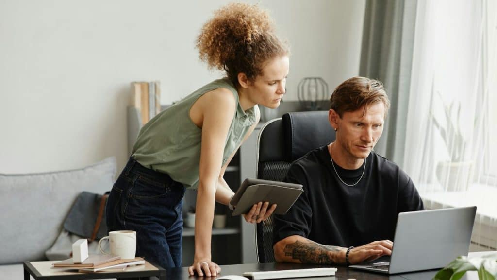 A man works on a laptop at a desk while a woman reviews a tablet nearby.