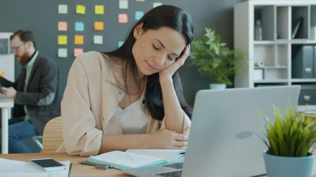 A woman sits at a desk, leaning on her hand with her eyes closed next to a laptop.