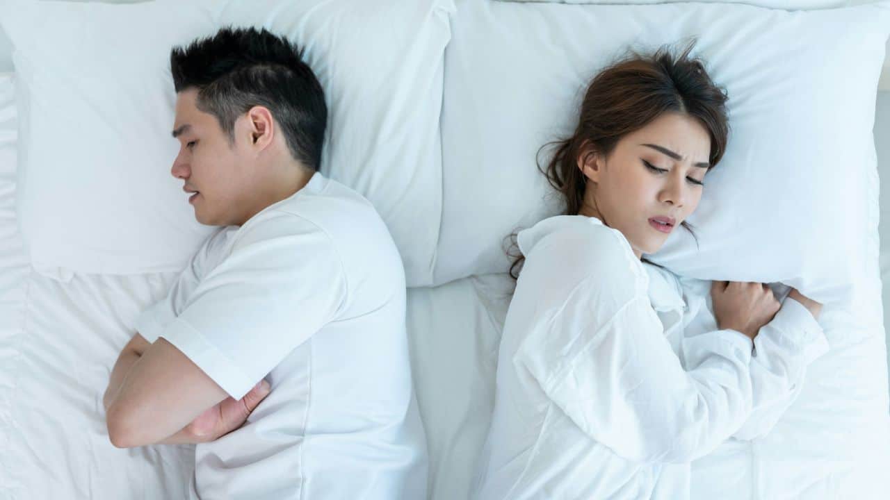 A man and woman lie in bed back-to-back with their arms crossed.