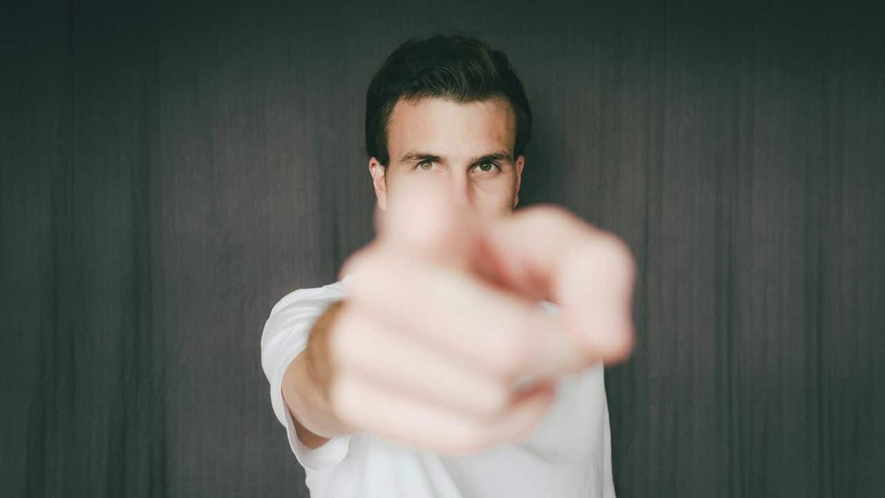 A man in a white shirt points directly at the camera with his hand blurred in the foreground.