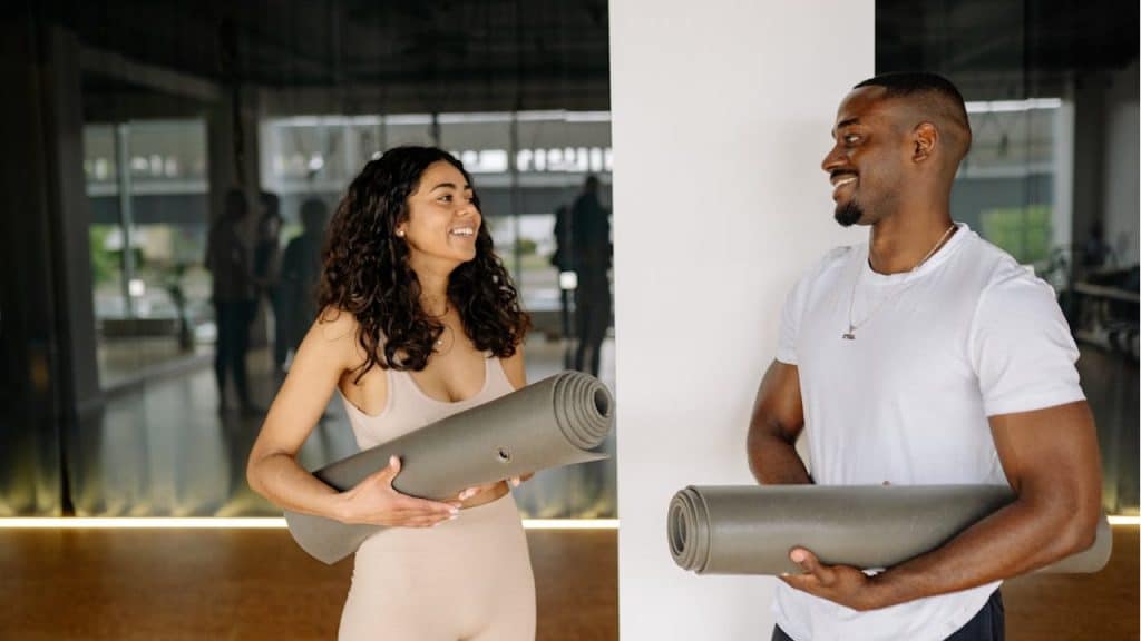A smiling man and woman are standing in a studio, both holding rolled-up yoga mats and looking at each other.