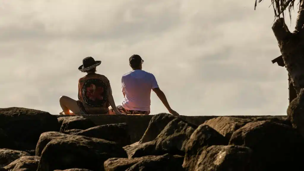 Two people sit on large rocks near the shore, facing away from the camera as they look out toward a cloudy sky.