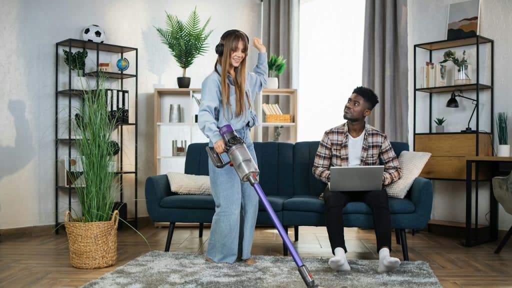 A woman vacuuming while a man sits on a couch and uses his laptop.