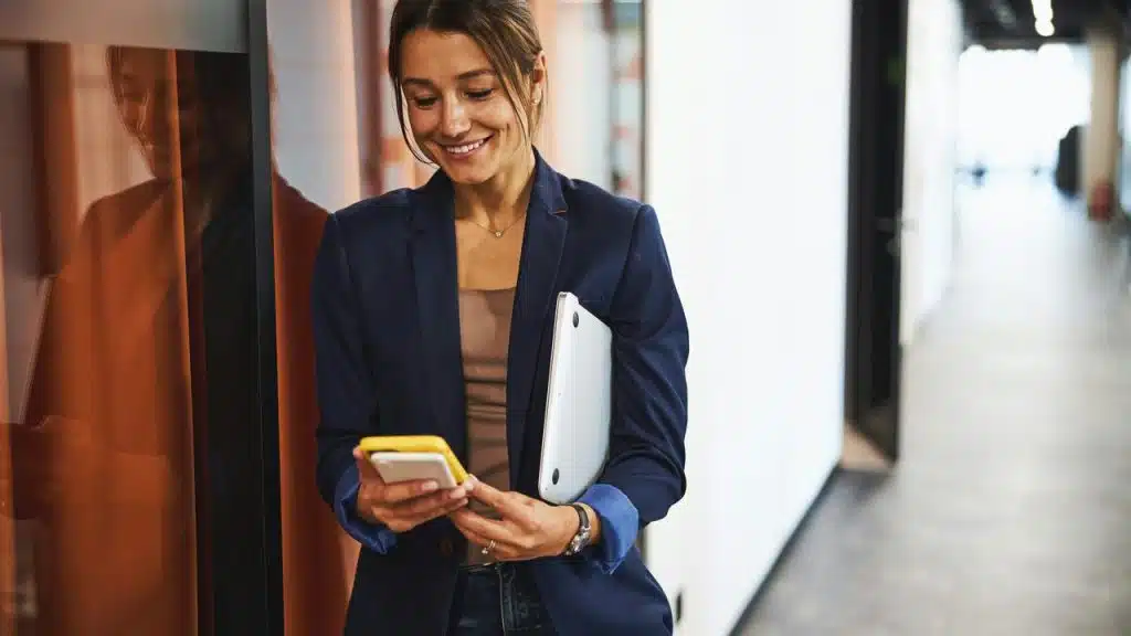 A woman in a blazer smiling at her phone while holding a laptop.