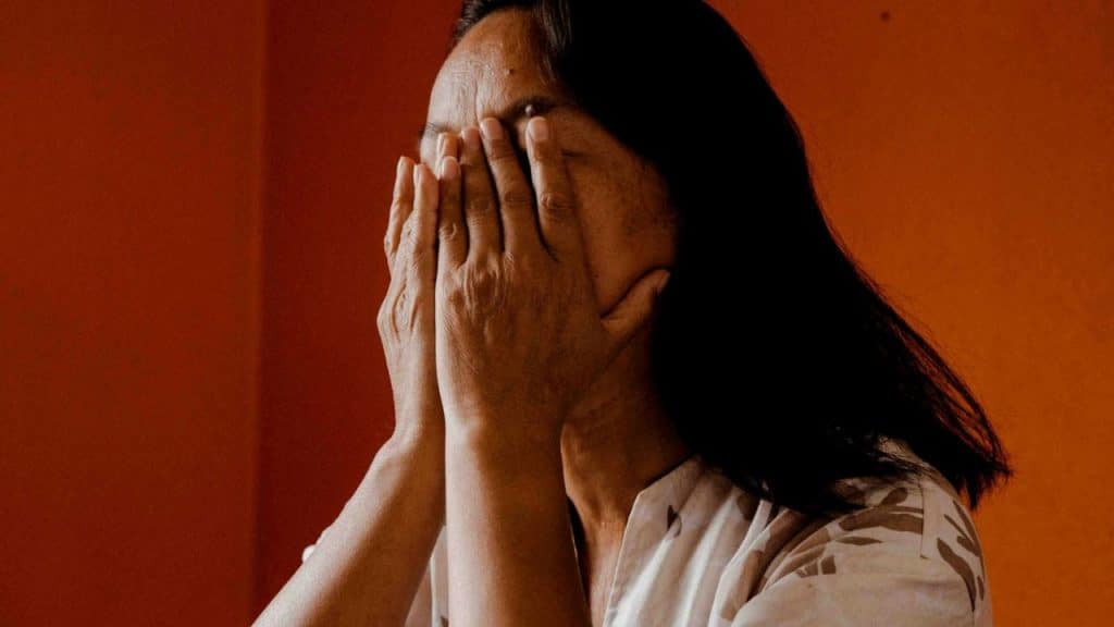 A person with long dark hair covers their face with both hands against an orange wall.