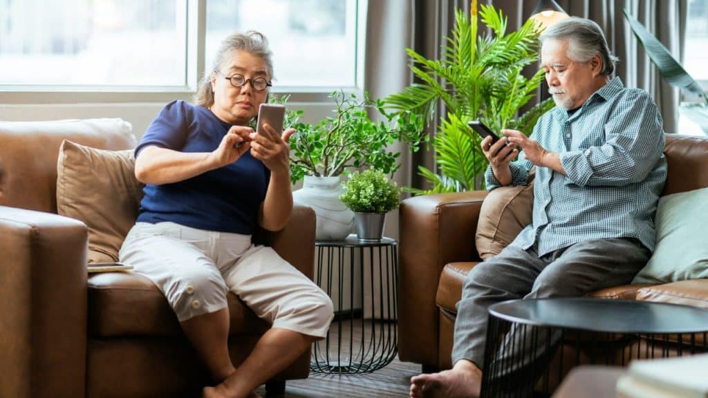 Two older adults sit separately on leather chairs, both looking down at their smartphones.