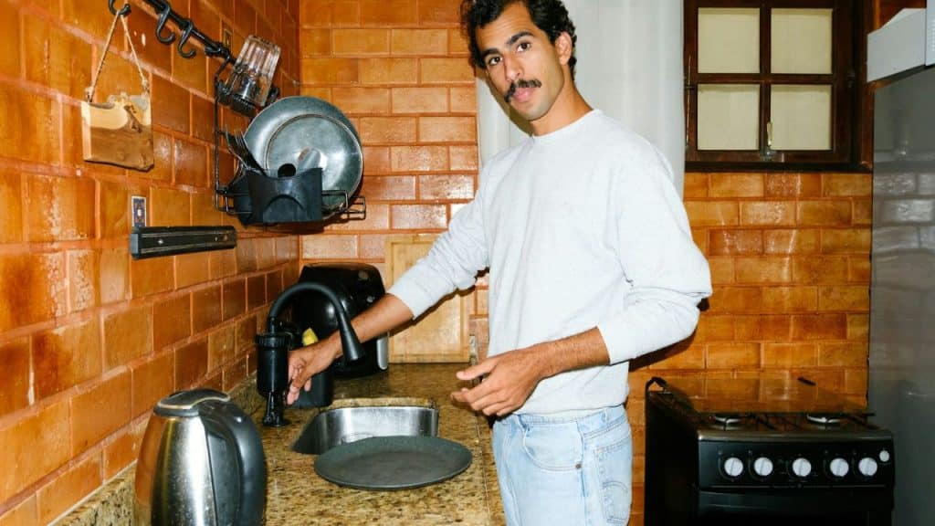 A man with a mustache in a light sweatshirt stands by a sink in a brick-walled kitchen.
