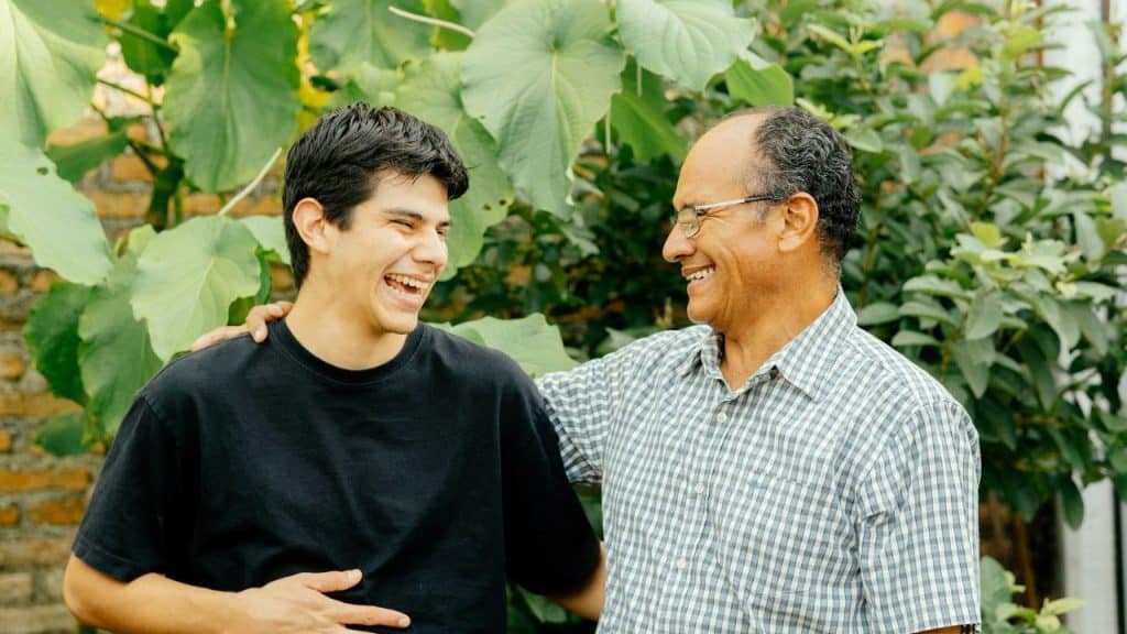 Father and son laughing outdoors with lush green plants in the background.