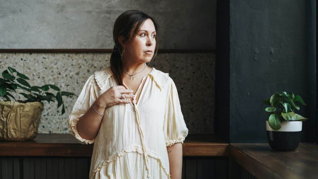 Woman in a loose beige dress standing by a window, looking away.