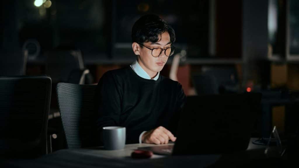 A man with glasses is sitting in a dark office, working on a laptop.