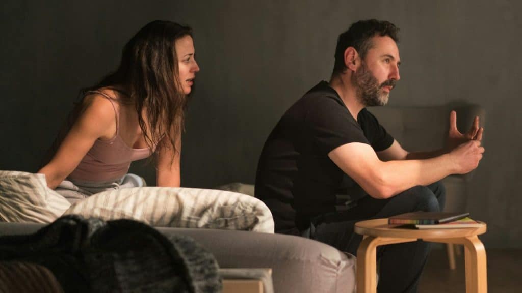 A man and woman sit next to each other in a dimly lit room, appearing tense.