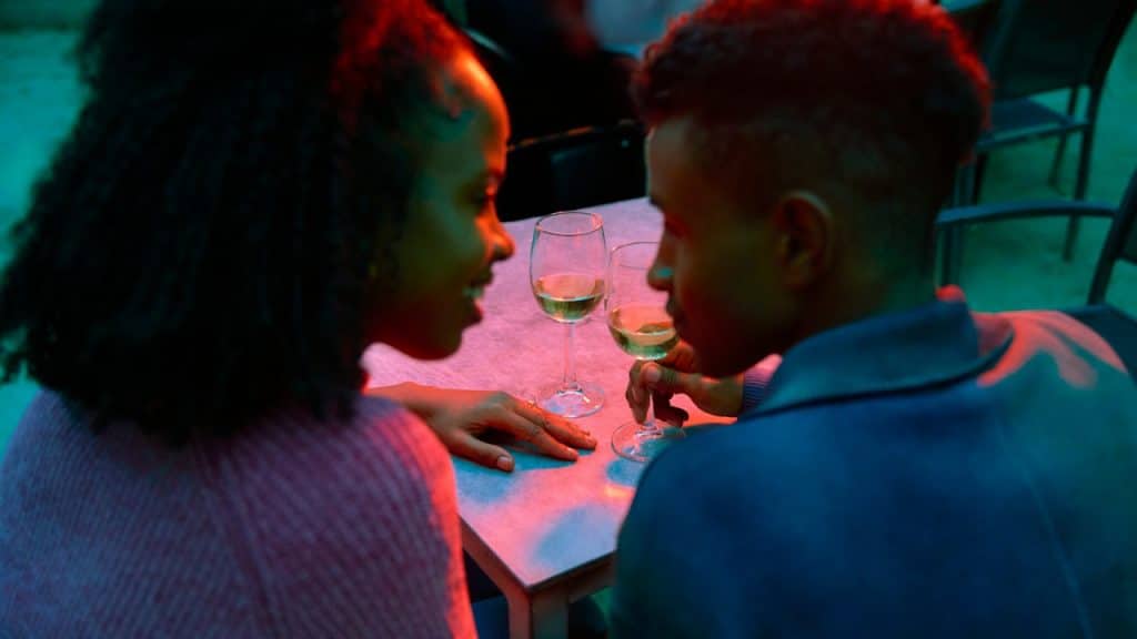 A couple is sitting across from each other at a table with wine glasses, illuminated by red and blue light, looking into each other's eyes.