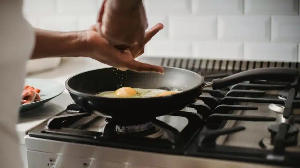 A person’s hand seasoning eggs cooking in a frying pan on a stovetop.