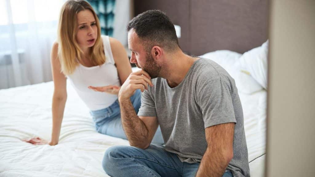 Woman arguing and gesturing at a man sitting with his hand on his chin.
