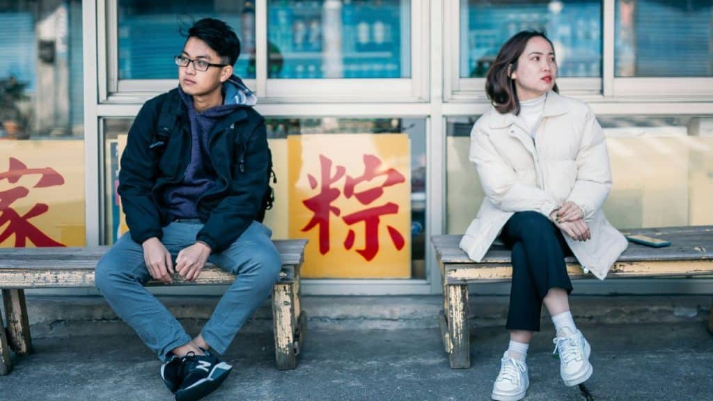 Two people sitting apart on wooden benches outdoors near a window with Chinese characters.