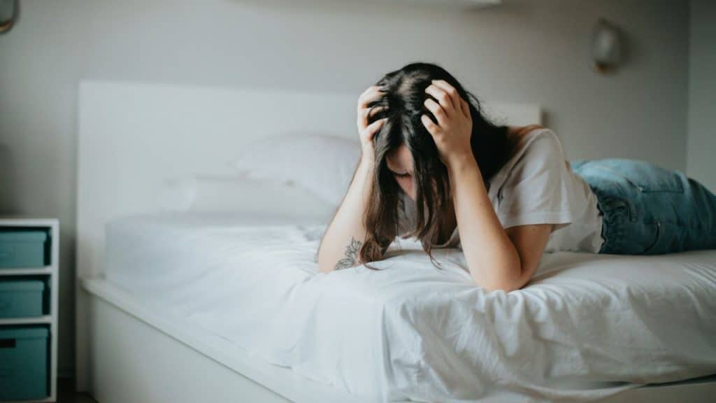 A woman is lying face down on a bed, holding her head with both hands.