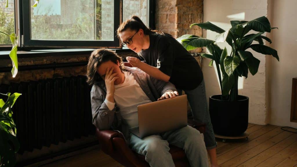 A stressed man covers his face while a woman talks to him over a laptop.