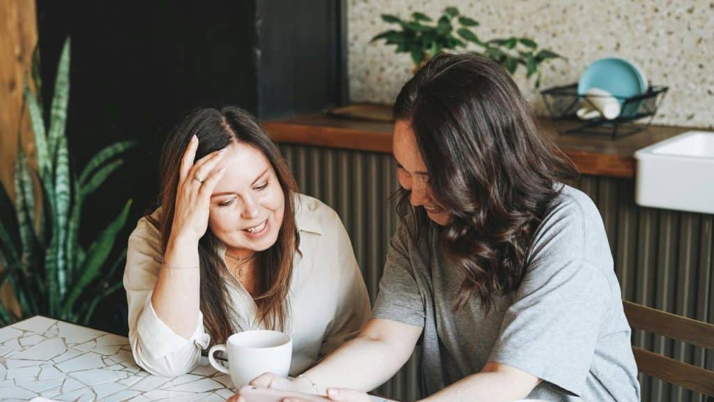 A photo of two women sitting at a table, smiling and looking at a smartphone together.
