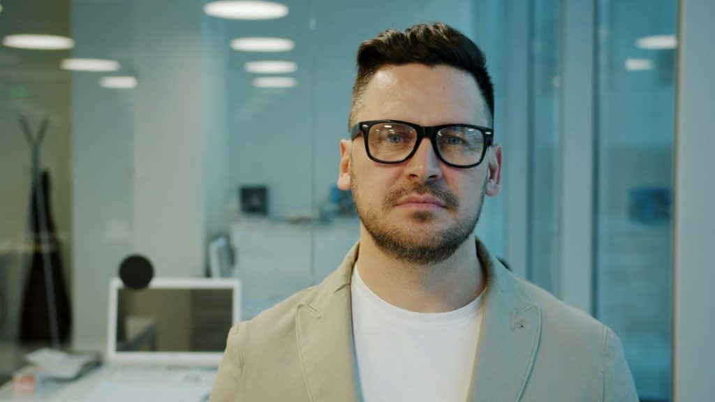 Close-up of a serious man in glasses and a light blazer in a modern office setting.