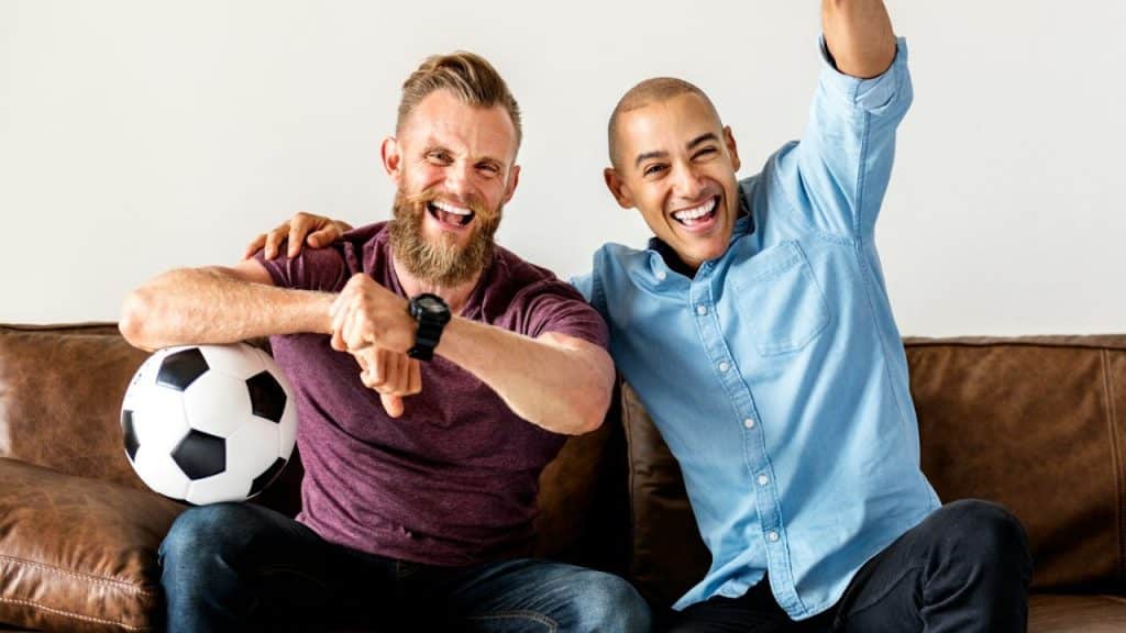 Two excited men cheering on a brown couch with a soccer ball, one bearded, the other raising his arm.