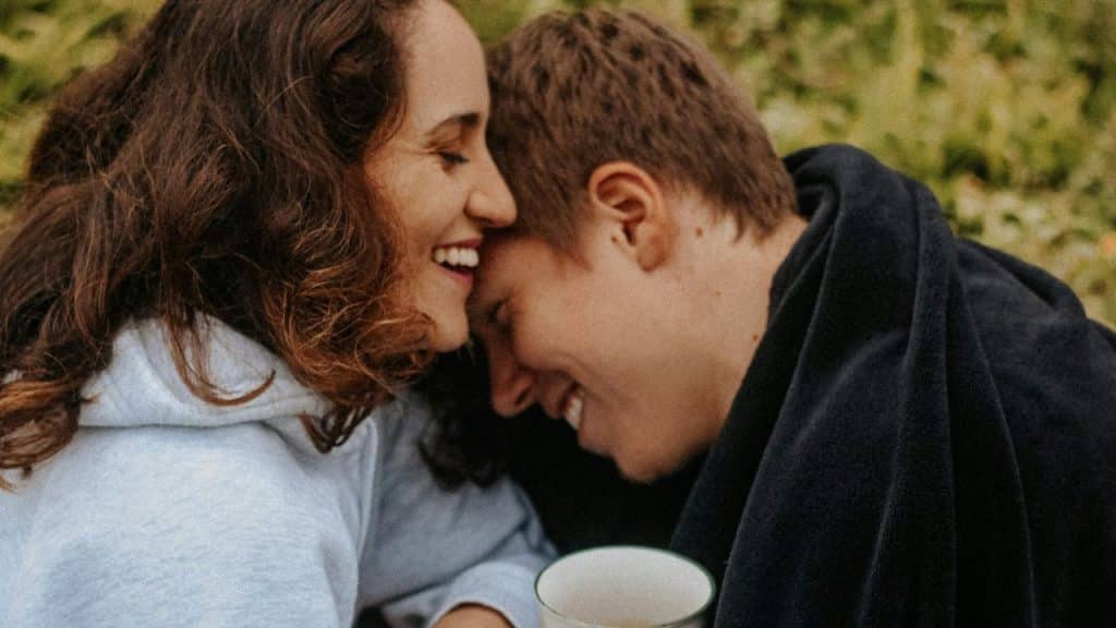 Two happy people with their heads together and smiling outdoors, with one covered by a blanket.