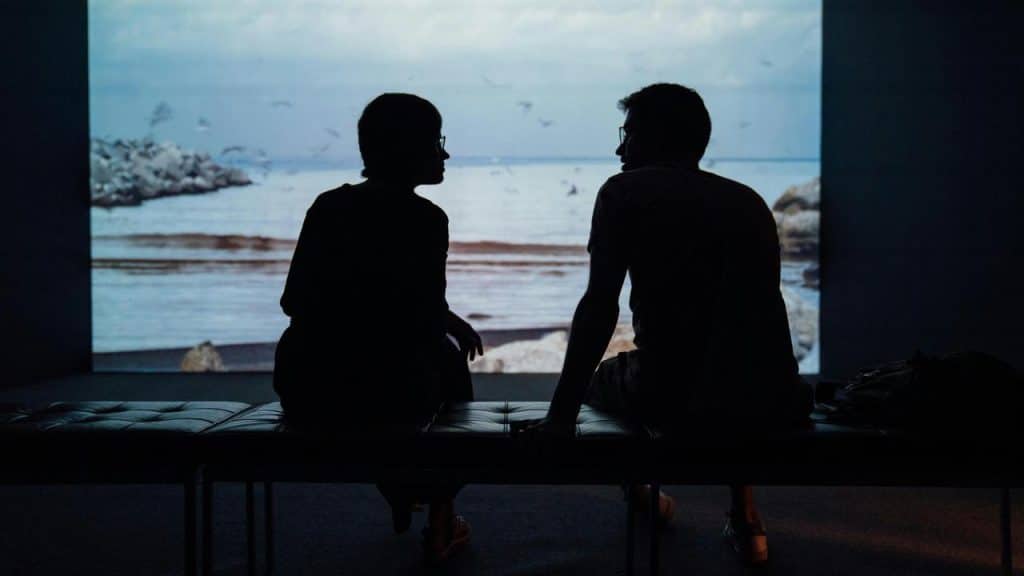 A silhouette of two people sitting and talking before a seascape screen.