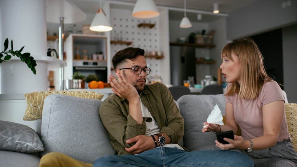 A man with annoyed expression sits on a couch while a woman beside him is talking intensely
