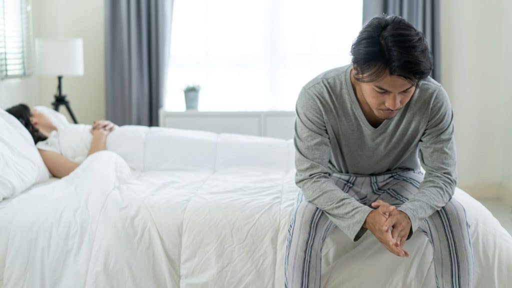 Man sitting sadly on a bed in foreground while a person looking in the window.