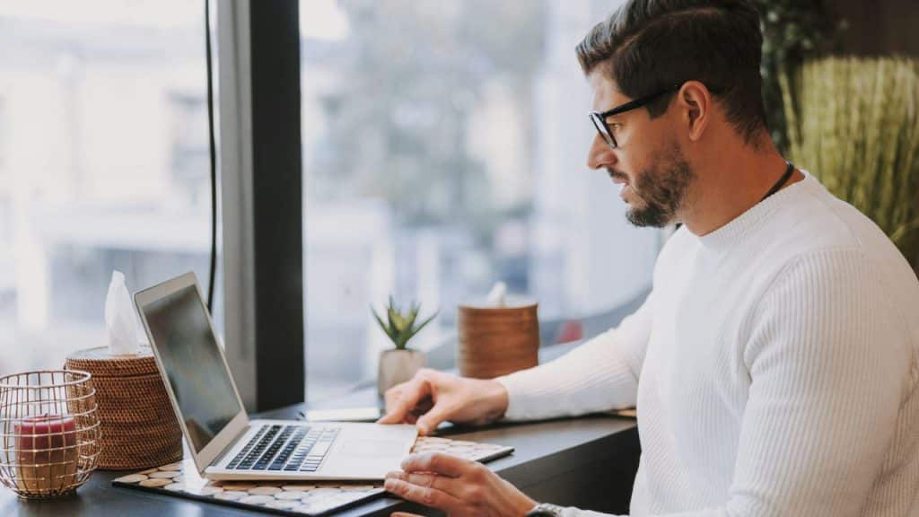 A bearded man wearing glasses and a white sweater working on a laptop.