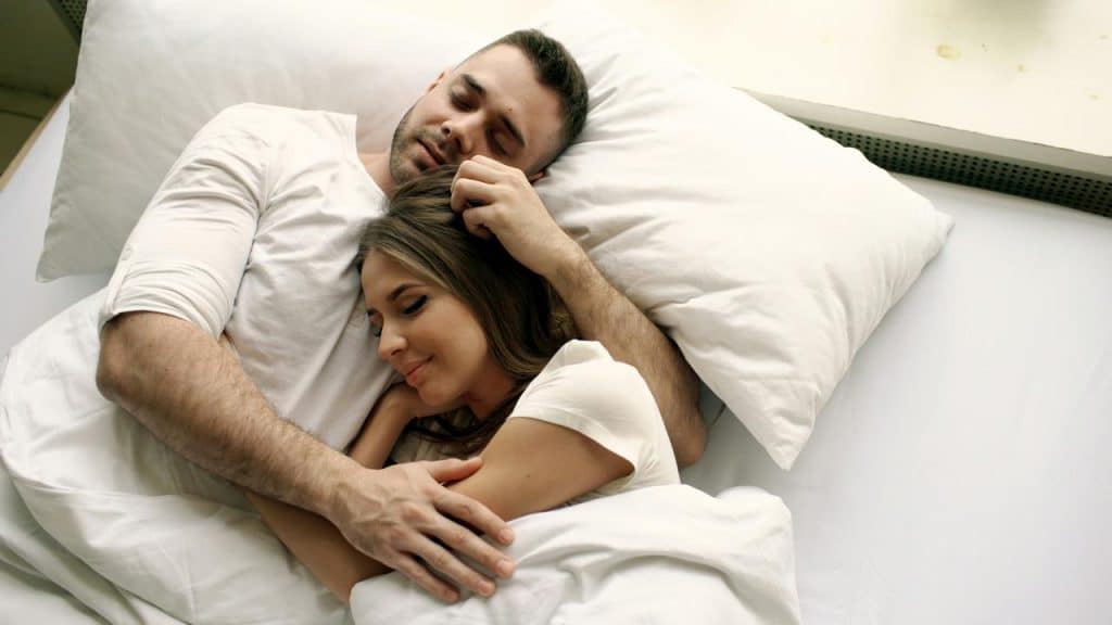 A couple cuddles together peacefully in bed, resting on white pillows and sheets.