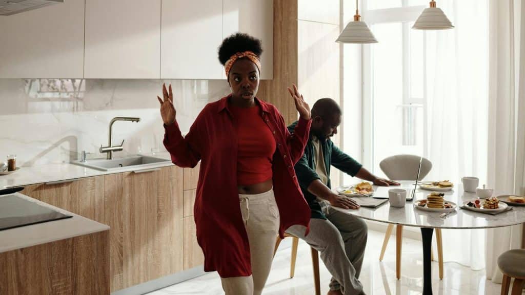 A woman standing in a kitchen with raised hands while a man sits at a table with food.