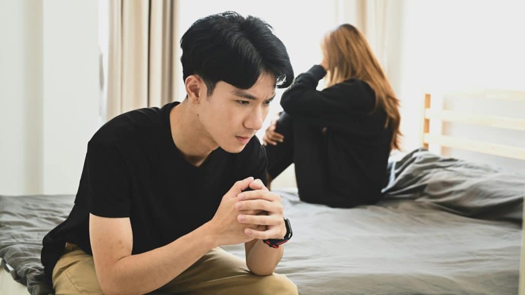 A Man looking depressed and sitting with crossed hands on a bed while a woman sits in the background.