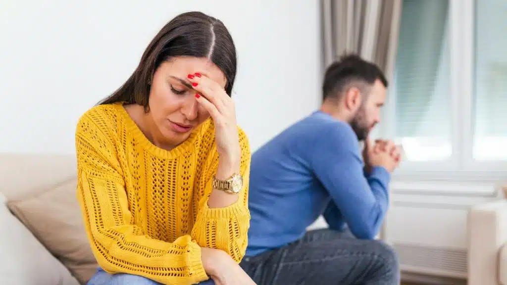 A distressed woman holds her forehead while a man sits hunched in the blurred background.
