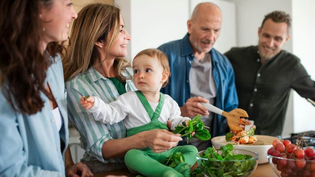 A happy multigenerational family indoors at home preparing vegetable salad in the kitchen.