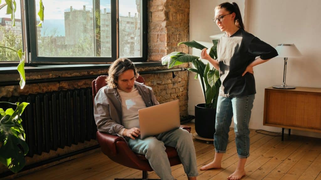 A man sitting while using his laptop being nagged by a woman wearing glasses.
