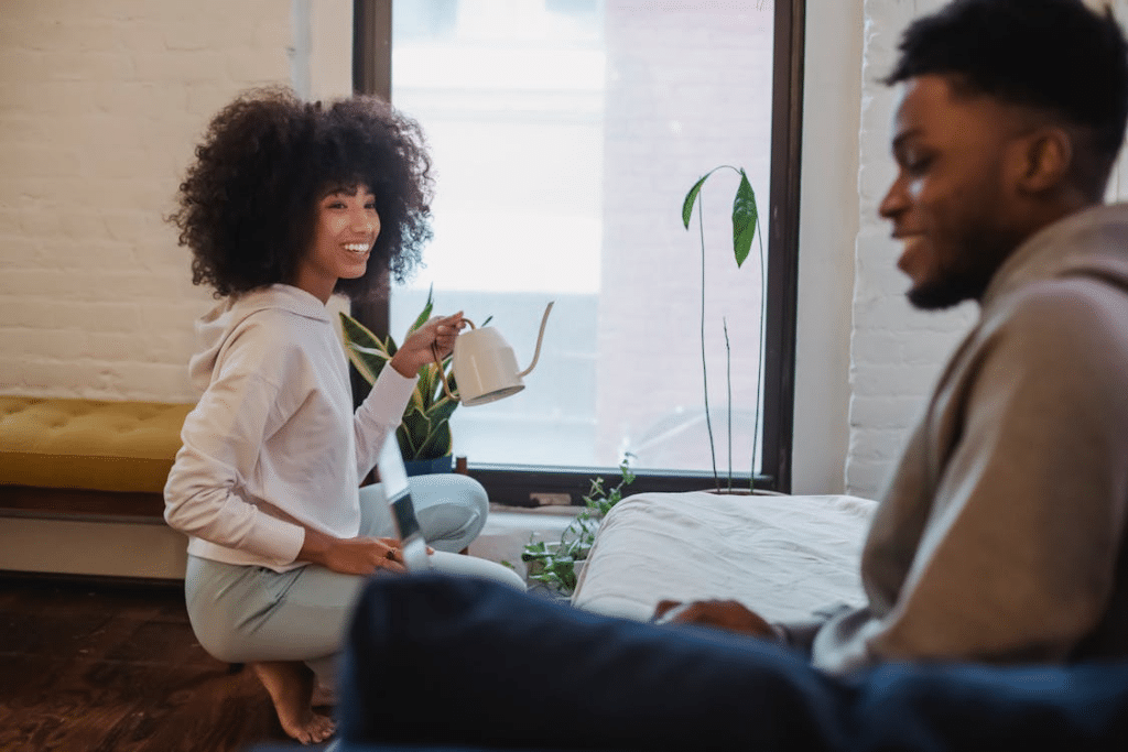 Positive black female watering flowers while spending time with companion at home