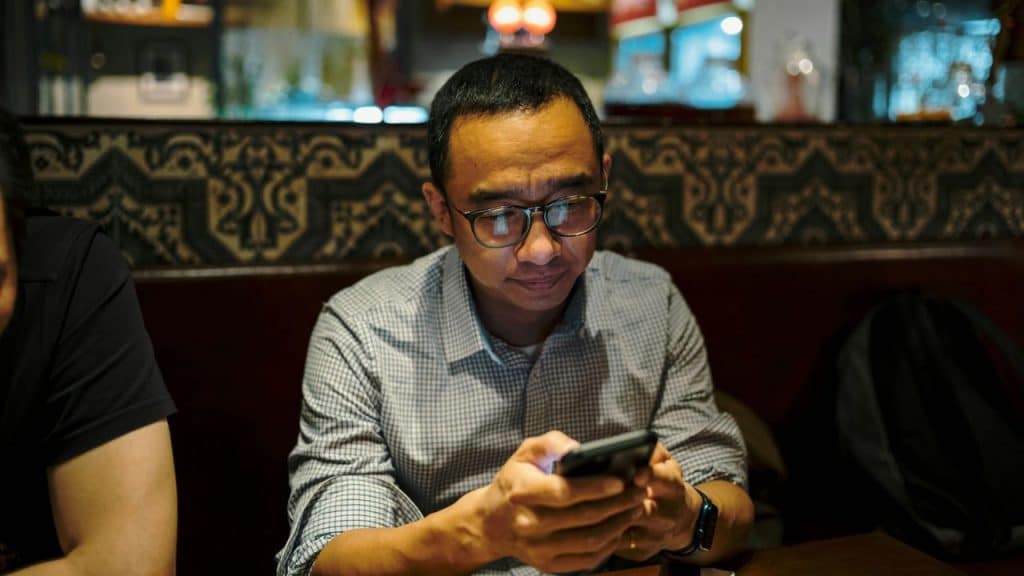 A man with glasses sits at a dark table looking intently at his phone.