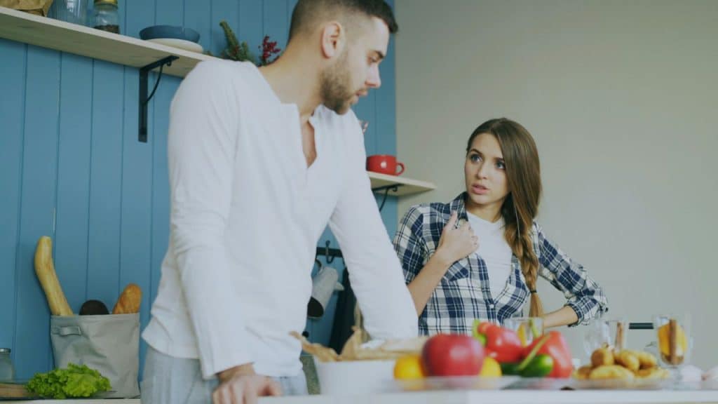 A man in a white shirt leans over a kitchen counter while a woman in a plaid shirt gestures while speaking to him.