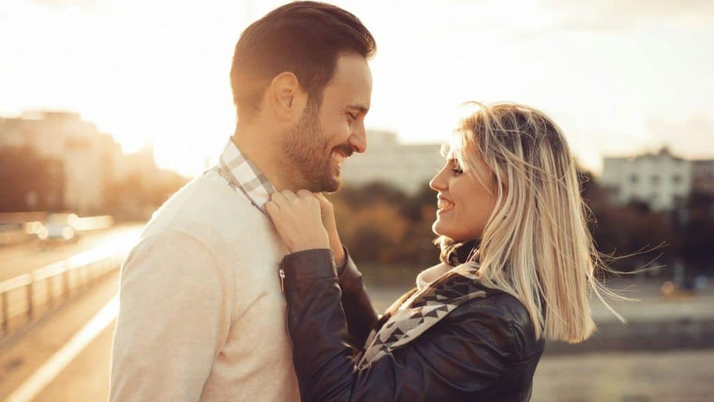 A smiling couple is looking at each other outdoors at sunset with a city in the background.