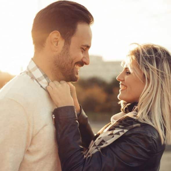 A smiling couple is looking at each other outdoors at sunset with a city in the background.
