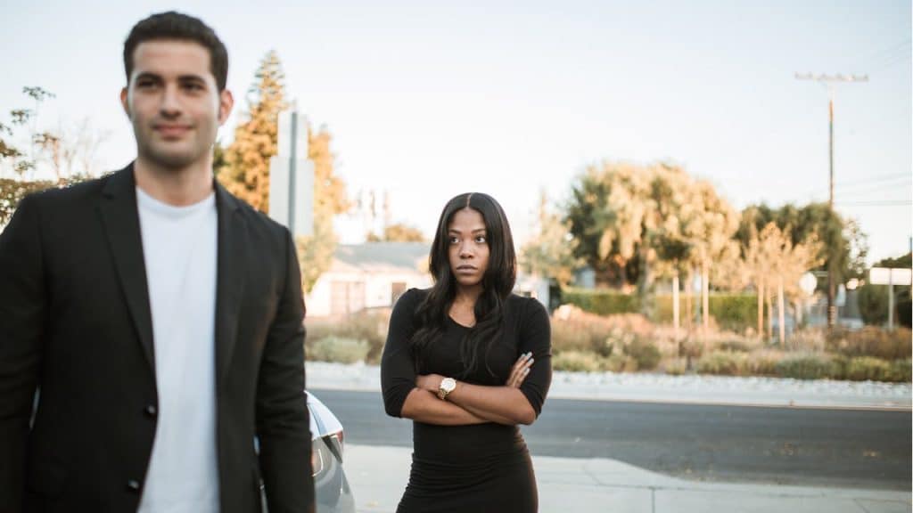 A serious-looking woman standing with her arms crossed and a slightly blurred man in a black jacket in the foreground.