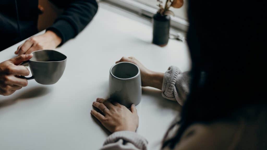 Overhead, close-up shot of two people's hands and arms, each holding a different-style gray coffee mug while seated at a light table by a window.