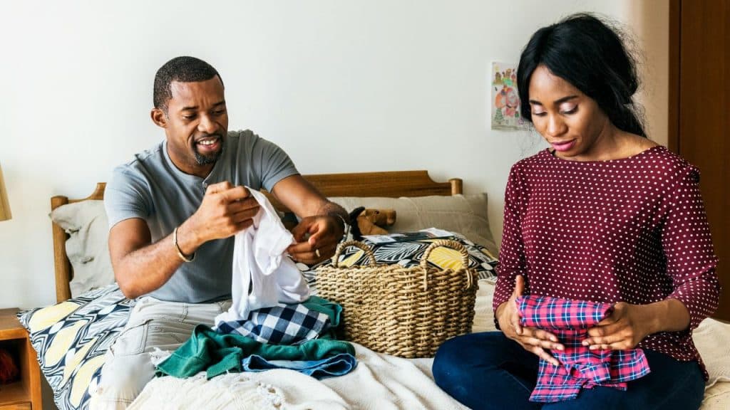 Man and woman sitting on a bed folding laundry from a woven basket.