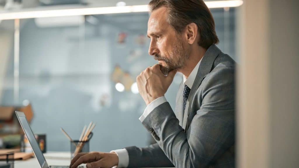 Focused businessman with a beard and suit working on a laptop with his chin resting on his fist.