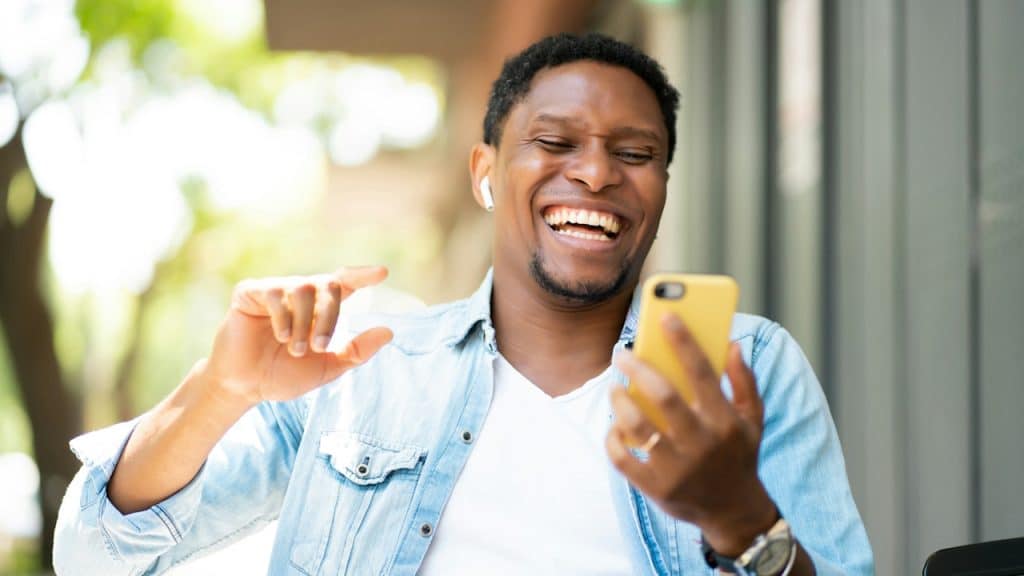 A man wearing a denim shirt, white t-shirt, and wireless earbuds is smiling and laughing while holding a yellow smartphone outdoors.