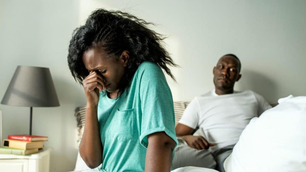 A distressed woman sits with her face in her hand while a man sits behind her.