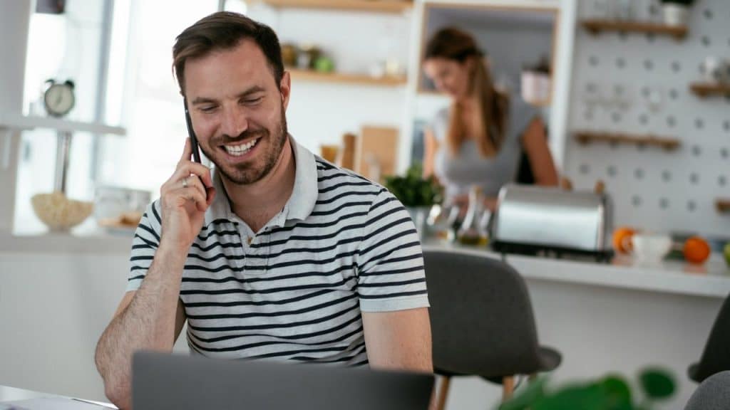 Smiling man in a striped shirt talking on a phone while working on a laptop in a bright kitchen.